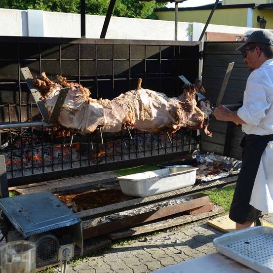 Ein Koch benutzt ein großes Messer, um ein ganzes gebratenes Schwein über einem Feuer zu zerlegen. Das Schwein befindet sich auf einem rotierenden Spieß mit einem Auffangbehälter darunter, um die Tropfen aufzufangen.