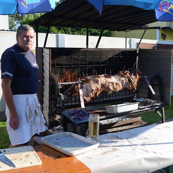 Ein Mann in einem blauen T-Shirt und einer weißen Schürze steht neben einem großen Outdoor-Grill mit einem gebratenen ganzen Schwein. Der Grill steht unter einem Vordach, mit einem Glas Bier und einem Tablett in der Nähe.