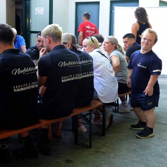 Mehrere Personen sitzen auf Bänken und tragen Uniformen mit der Aufschrift 'Neufelder'. Ein Mann steht in der Nähe und lächelt, er trägt ein blaues Shirt. Andere sind im Hintergrund zu sehen, stehend in der Nähe von Türen.