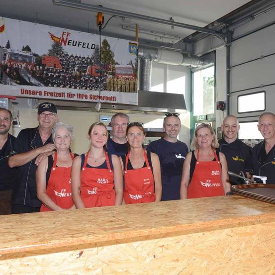 Eine Gruppe von Menschen in roten Schürzen steht vor einem Tresen, wahrscheinlich in einer Küche. Ein Banner hinter ihnen trägt die Aufschrift 'FF NEUFELD' und zeigt eine Gruppe von Feuerwehrleuten.