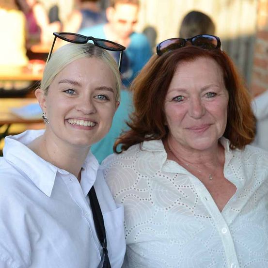 Zwei Frauen posieren für ein Foto bei einer Outdoor-Veranstaltung. Eine hat kurze blonde Haare und eine Brille, die andere hat rote Haare und eine Halskette. Sie tragen beide eine Sonnenbrille. Verschwommene Personen sind im Hintergrund zu sehen.