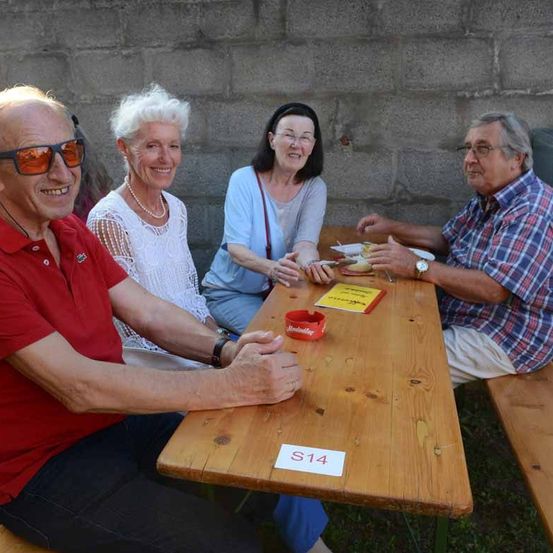 Vier ältere Erwachsene sitzen um einen holzernen Tisch mit der Aufschrift S14 und posieren für ein Foto. Sie sind draußen in der Nähe einer Steinmauer.