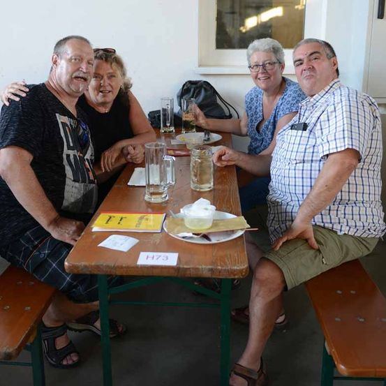 Vier Personen sitzen an einem Tisch mit Getränken und Essen. Zwei Männer und zwei Frauen, alle lächeln. Der Mann rechts hält ein Bierglas. Der Tisch hat einen Teller mit Essen und ein gelbes Schild.