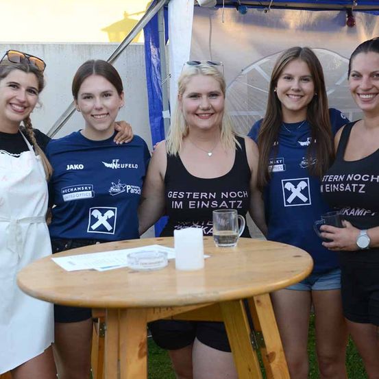 Fünf Frauen posieren bei einer Outdoor-Veranstaltung zusammen und halten Getränke. Sie tragen passende blaue T-Shirts mit weißen Texten und Logos. Die Frau in der Mitte hält ein Glas, während andere für die Kamera lächeln. Ein Holztisch und ein Zelt sind im Hintergrund.