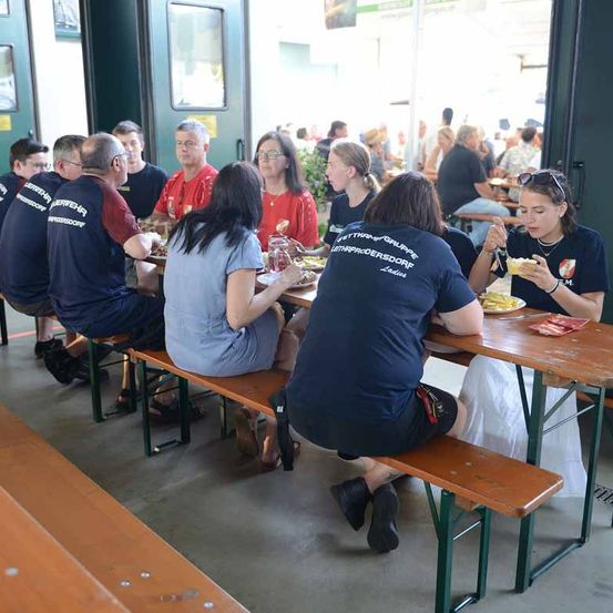 Eine Gruppe von Menschen isst in einem Restaurant. Sie sitzen auf Holzbänken um einen Tisch herum. Sie tragen Freizeitkleidung. Einige von ihnen tragen Brillen.