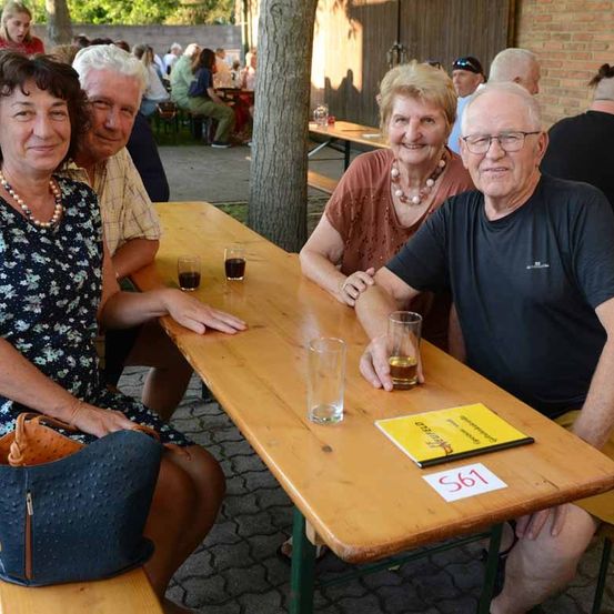 Vier ältere Menschen sitzen auf einer Bank bei einer Veranstaltung im Freien. Sie lächeln, mit Getränken und der Nummer 561 auf dem Tisch.