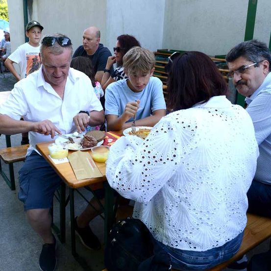 Eine Gruppe von Menschen isst im Freien. Ein älterer Mann schneidet Essen. Eine Frau davor hat ein gemustertes Oberteil. Ein Junge neben ihr schaut nach unten. Die anderen sitzen hinter ihnen.