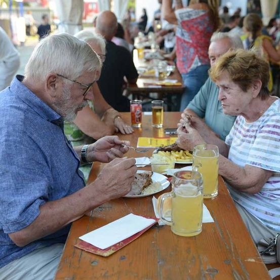 Eine Szene des Essens im Freien mit älteren Menschen, die an einem langen Tisch sitzen, essen und trinken. Ein Mann mit Brille hält eine Gabel, und eine Frau neben ihm trägt ein gestreiftes Hemd. Sie haben Teller mit Essen und Gläser mit Getränken. Im Hintergrund sitzen andere Menschen an Tischen, und eine Frau steht.