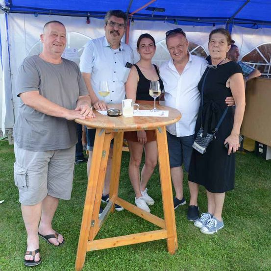 Fünf Personen stehen unter einem blauen Zelt und lächeln für ein Foto. Eine Frau in Schwarz hält den Arm eines Mannes. Ein Tisch hat zwei Weingläser, eine Tasse und einen Teller.