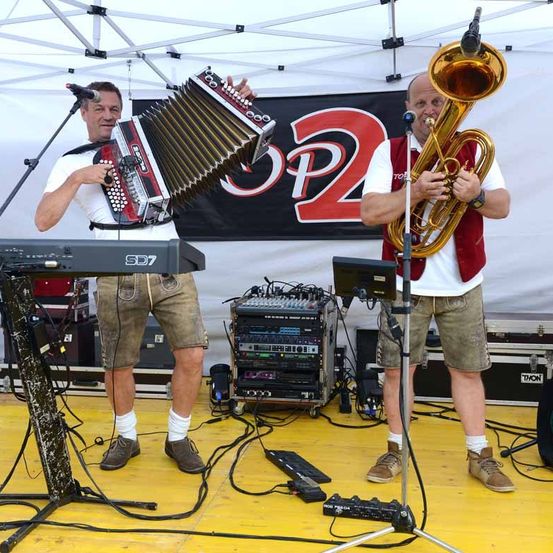 Zwei Männer spielen auf der Bühne mit Mikrofonen. Einer spielt Akkordeon und der andere Tuba. Hinter ihnen befindet sich ein Banner mit dem Text 'SP2'.