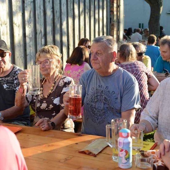 Mehrere ältere Personen sitzen um einen Holztisch herum und heben Gläser mit dunklem Bier. Der Hintergrund zeigt eine verschwommene Menschenmenge, möglicherweise in einem Garten oder bei einer Outdoor-Veranstaltung.