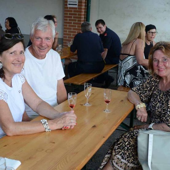 Zwei Frauen sitzen an einem Tisch mit Weingläsern und einem Mann in einem weißen T-Shirt. Dahinter steht eine Ziegelmauer und Leute sitzen an Bänken. Ein Schild heißt Hopfburg.