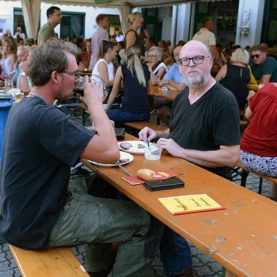 Zwei Männer sitzen an einem Tisch in einem überfüllten Outdoor-Restaurant, beide mit Brille. Ein Mann trinkt aus einem Glas, während der andere Essen von einem Teller isst.