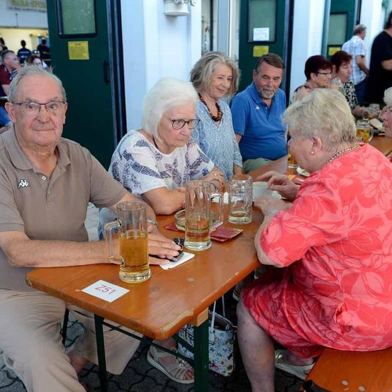 Eine Gruppe älterer Erwachsener sitzt um einen Holztisch herum und trinkt Bier. Sie tragen lässige Sommerkleidung. Eine Frau in einem rosa Kleid schaut den Mann im braunen Hemd an.