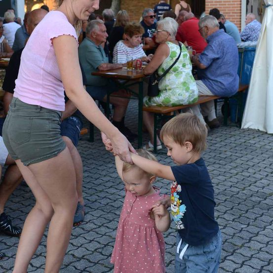 Eine Frau hält die Hände zweier Kinder, die sich an den Händen halten, bei einer Outdoor-Veranstaltung. Viele Menschen sitzen an Tischen, einige stehen.