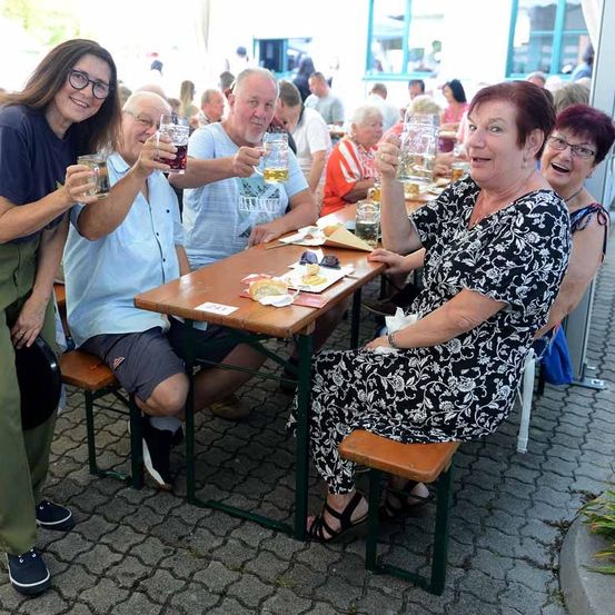 Eine Gruppe von Menschen, darunter Männer und Frauen, sitzt an einem Tisch und hält Gläser mit Bier in der Hand. Sie lächeln und scheinen sich bei einer Veranstaltung im Freien zu amüsieren.
