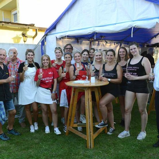 Eine Gruppe von Frauen und Männern posiert für ein Foto vor einem Tisch mit Weingläsern unter einem Zelt. Sie tragen passende schwarze Tanktops mit unterschiedlichen Slogans.