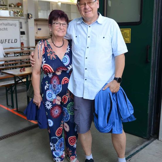 Ein Mann und eine Frau stehen zusammen in einem Raum mit einer grünen Tür. Die Frau trägt ein Blumenkleid und der Mann hält eine blaue Jacke. Sie lächeln beide. Hinter ihnen steht ein Tisch mit Bänken und einem Regal mit verschiedenen Gegenständen.