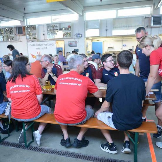 Mehrere Personen sind in einem Raum versammelt, einige sitzen auf Bänken und andere stehen. Sie tragen rote und blaue T-Shirts mit den Worten 'Feuerwehr' und 'Lettland'. Eine Frau steht zwischen zwei Männern. Ein Banner mit den Worten 'Gute Unterhaltung wünscht' ist an der Wand angebracht.