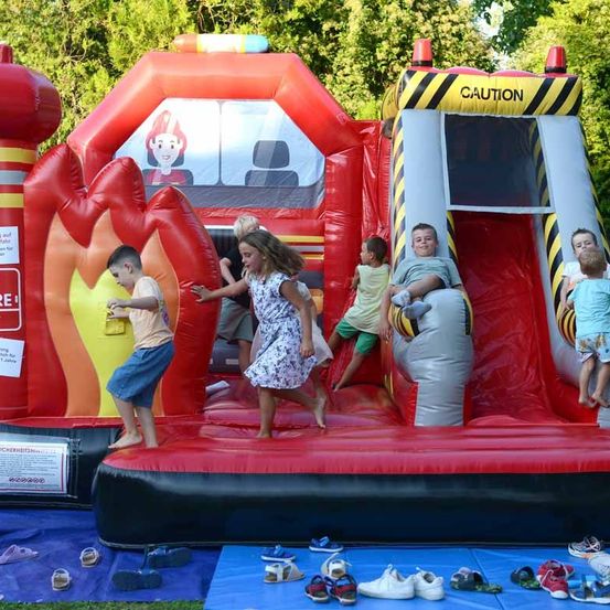 Mehrere Kinder spielen auf einem großen aufblasbaren Feuerwehrauto. Ein Mädchen und ein Junge rutschen herunter, während andere stehen und draußen spielen. Schuhe sind auf einem blauen Tarp verstreut.