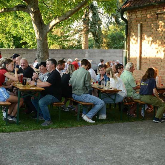 Eine Gruppe von Menschen sitzt an Tischen vor einem Gebäude. Sie trinken und scheinen zu plaudern. Die Tische sind unter einem Baum aufgebaut.