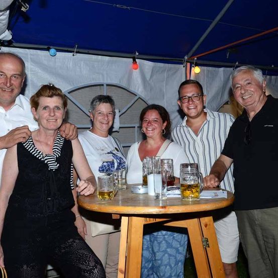 Fünf Menschen lächeln unter einem blauen Zelt mit Gläsern Bier auf einem kleinen Tisch.