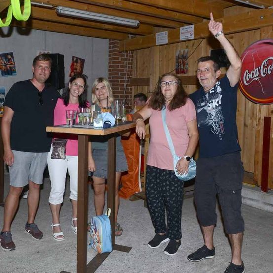 Eine Gruppe Erwachsener steht um einen kleinen Tisch mit Getränken in Gläsern herum. Dahinter ist ein Coca-Cola-Schild an einer Holzwand zu sehen. Die Umgebung wirkt leger und gesellig.