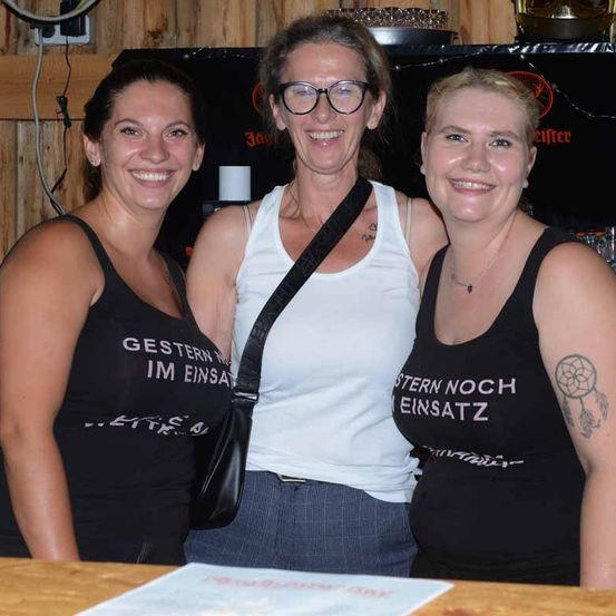 Drei Frauen stehen lächelnd zusammen in einer Bar. Die Frau in der Mitte trägt eine Brille und ein weißes Tanktop. Die anderen tragen schwarze Tanktops mit weißem Text. Eine Holzwand und eine schwarze Bartheke sind im Hintergrund.