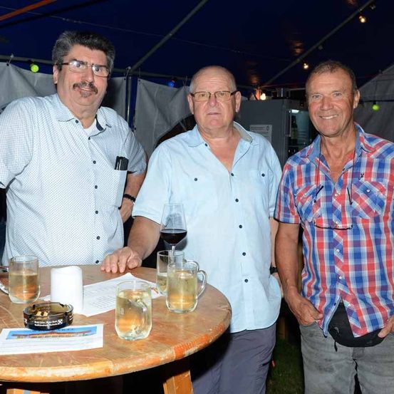 Drei Männer stehen neben einem Tisch mit Gläsern Wein und Wasser unter einem Zelt. Der Mann auf der linken Seite trägt ein weißes Hemd, der in der Mitte trägt ein blaues Hemd und der auf der rechten Seite trägt ein rot-blau kariertes Hemd.