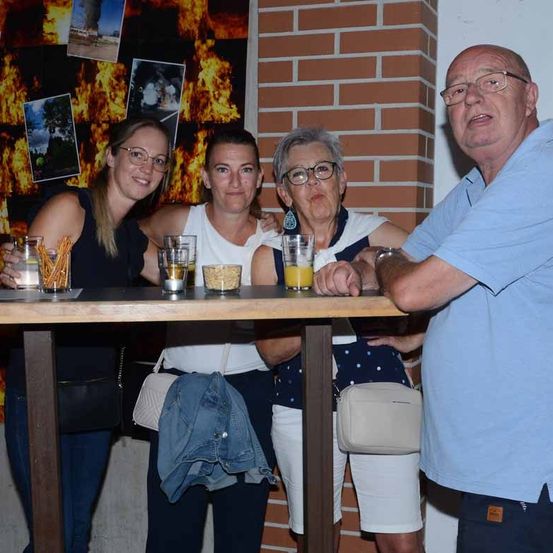 Vier Personen stehen hinter einem Tisch mit Getränken, darunter ein Glas Orangensaft. Hinter ihnen befindet sich eine Ziegelwand mit Feuerbildern.