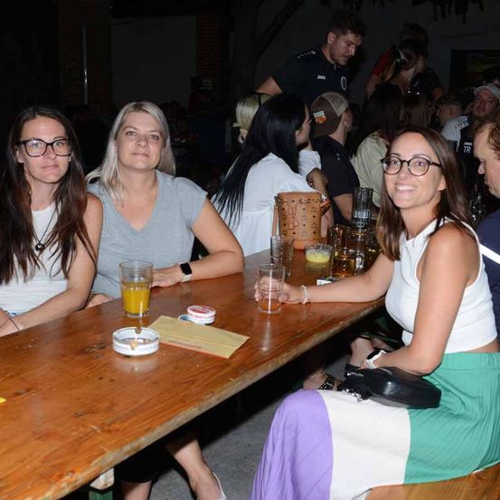 Eine Gruppe von Frauen sitzt zusammen an einem Tisch und lächelt. Der Tisch ist mit Gläsern und Aschenbechern gefüllt. Ein Sicherheitsbeamter steht hinter ihnen.