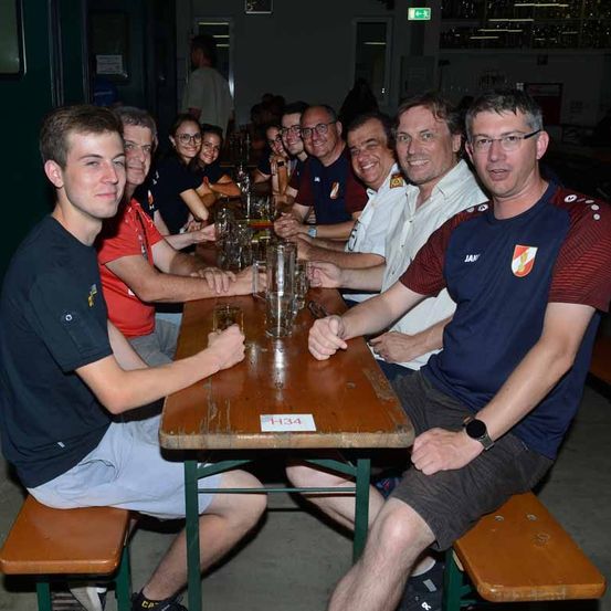 Eine Gruppe von Erwachsenen in Freizeitkleidung, die um einen Holztisch mit Gläsern sitzen, lächeln und für ein Foto posieren.