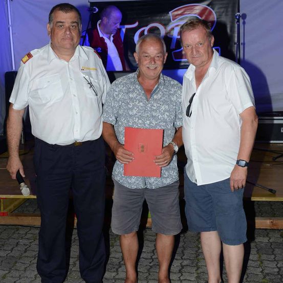 Drei Männer stehen zusammen; der mittlere Mann hält eine rote Mappe. Ein Mann links trägt eine Militäruniform und ein anderer eine Uhr. Ein Banner steht hinter ihnen, möglicherweise bei einer Veranstaltung.