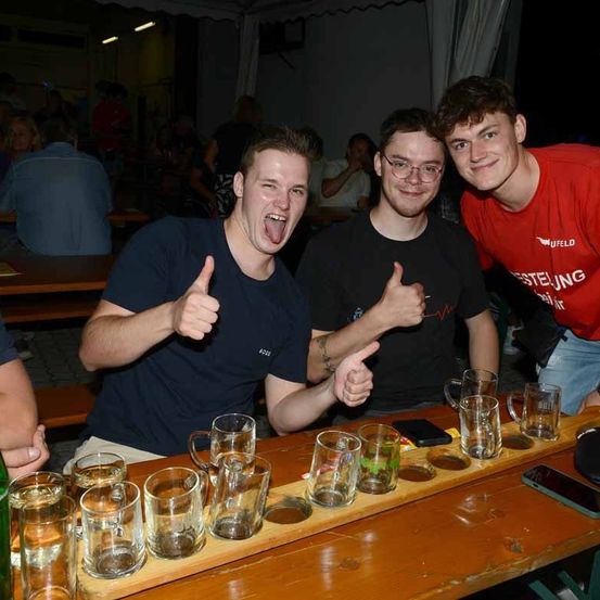 Drei junge Männer zeigen Daumen hoch vor einer Bar, Gläser mit Bier stehen auf einem Tablett vor ihnen.