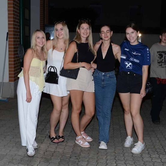 Fünf Frauen in verschiedenen Outfits stehen nachts zusammen draußen. Eine trägt ein gelbes Oberteil, eine andere ein schwarzes Oberteil und Jeansshorts. Hinter ihnen steht ein Ziegelgebäude.