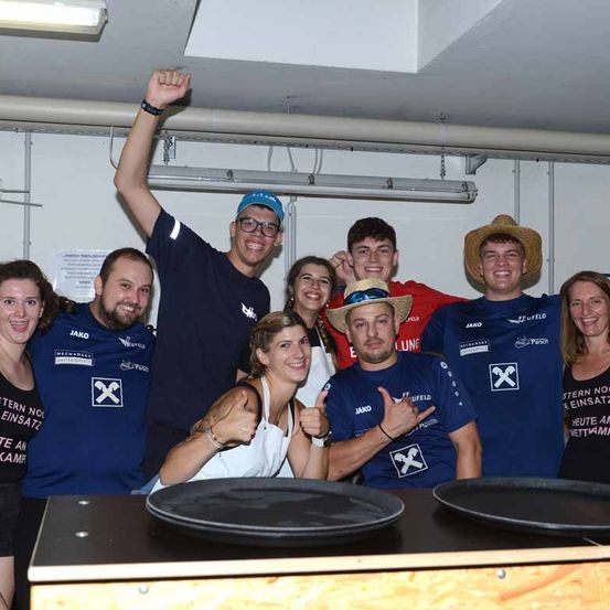 Eine Gruppe von Menschen in blauen T-Shirts posiert für ein Foto in einem Raum mit weißen Wänden und Beleuchtungskörpern. Sie lächeln und machen verschiedene Gesten.