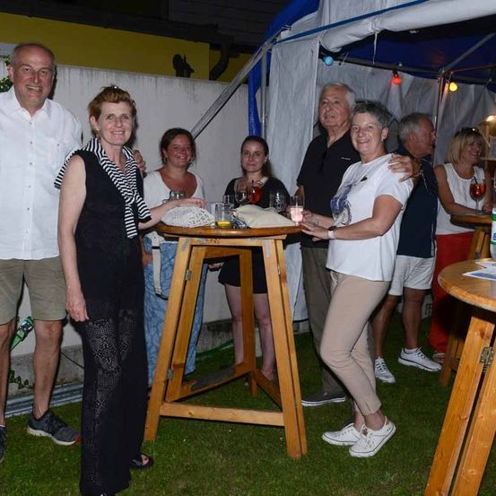 Eine Gruppe von Menschen steht um einen kleinen Tisch herum, hält Getränke und lächelt. Eine Frau hält ein gestricktes Objekt. Hinter ihnen steht ein weißes Zelt und eine gelbe Wand.