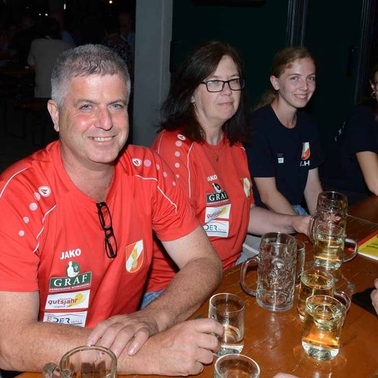 Drei Personen sitzen an einem Tisch mit Biergläsern, alle tragen rote T-Shirts. Der Mann trägt eine Brille und lächelt, während die Frauen zusehen. Im Hintergrund sitzen weitere Personen an einem Tisch.