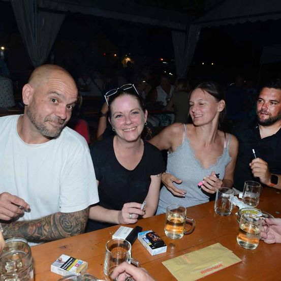 Eine Gruppe von Menschen sitzt an einem Tisch mit Getränken. Ein Mann auf der linken Seite hat Tattoos auf seinem Arm. Eine Frau in der Mitte hat eine Brille auf dem Kopf. Eine andere Frau auf der rechten Seite lächelt. Ein Mann neben ihr hat einen Stift. Sie sind in einer Bar.