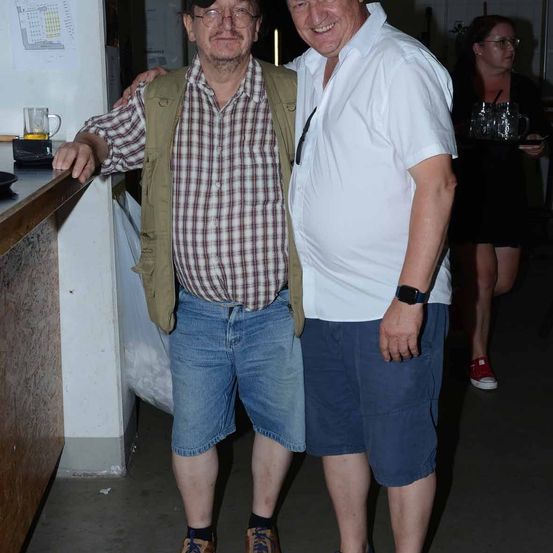 Zwei Männer stehen zusammen und lächeln für ein Foto. Der Mann auf der linken Seite trägt ein kariertes Hemd und eine Weste, während der Mann auf der rechten Seite ein weißes Hemd und blaue Shorts trägt. Eine Frau geht hinter ihnen und trägt ein Tablett mit Gläsern.