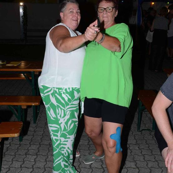 Zwei Frauen tanzen bei einer Outdoor-Veranstaltung bei Nacht. Eine trägt ein weißes Tanktop, die andere ein grünes Hemd und schwarze Shorts. Im Hintergrund unterhalten sich Leute an Tischen.