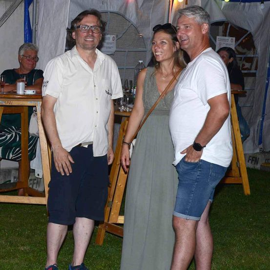 Drei Menschen stehen draußen in der Nähe eines Zeltes. Eine Frau steht zwischen zwei Männern. Sie scheinen zu lächeln und posieren für ein Foto.