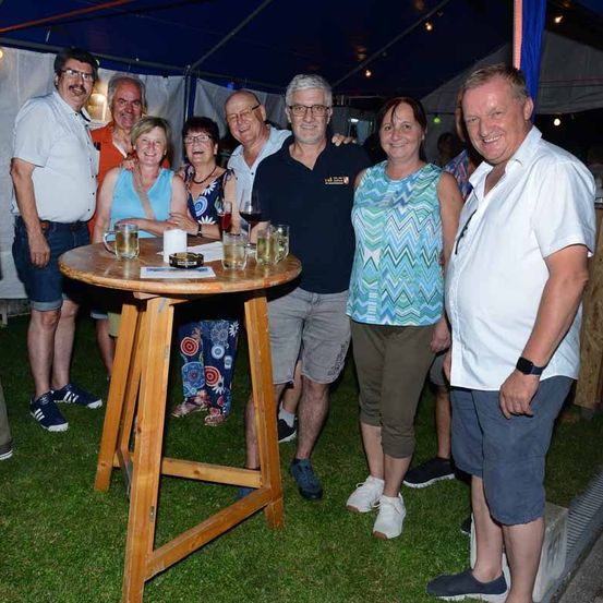 Eine Gruppe von Menschen steht draußen in einem Zelt und lächelt für ein Foto. Sie sind bei einem Tisch mit Biergläsern, einer Kerze und anderen Gegenständen. Ein Mann hält ein Glas Wein.