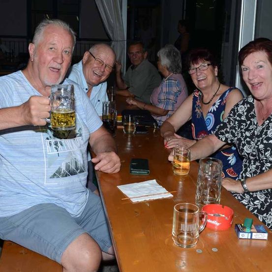 Eine Gruppe von Menschen sitzt an einem Tisch, einige halten Biergläser. Sie scheinen einen Abend zu genießen. Auf dem Tisch befinden sich verschiedene Gegenstände, darunter Handys, eine Geldbörse und eine Schachtel Zigaretten. Die Umgebung scheint sich im Innenbereich zu befinden.