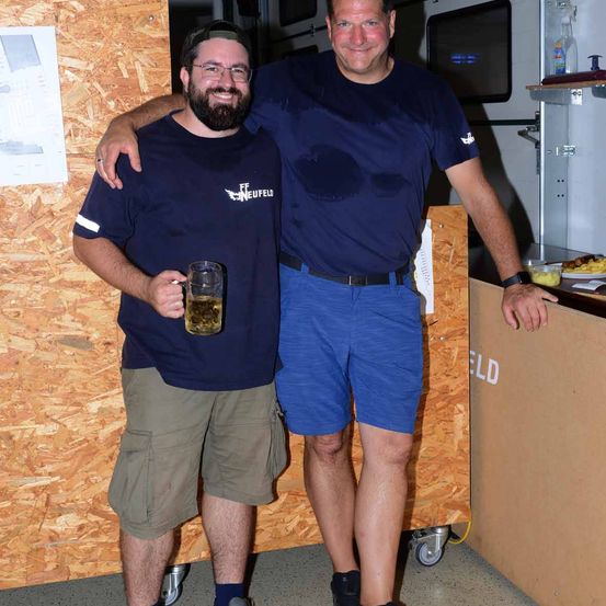 Zwei Männer in identischen blauen T-Shirts, einer mit einem Bierkrug, posieren für ein Foto in einer Küchenbereich mit Holzwänden und einer Theke.