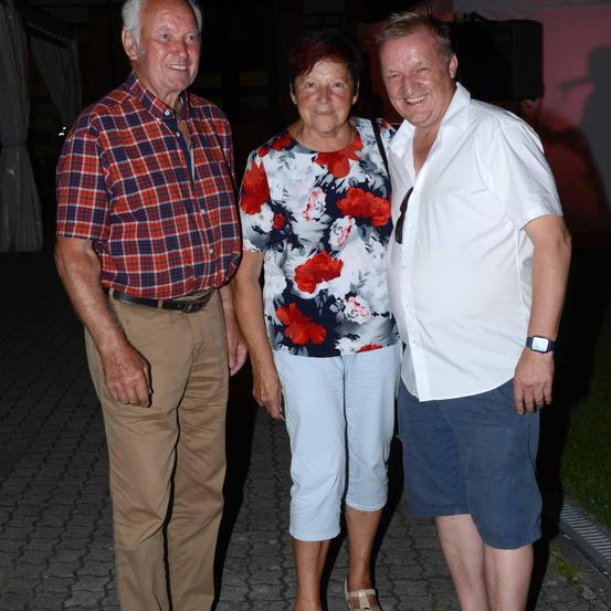 Drei Erwachsene stehen bei Nacht auf einem Gehweg aus Kopfsteinpflaster. Der Mann links trägt ein kariertes Hemd und Khakihose. Die Frau in der Mitte trägt ein Blumenoberteil und weiße Shorts. Der Mann rechts trägt ein weißes Hemd und blaue Shorts.