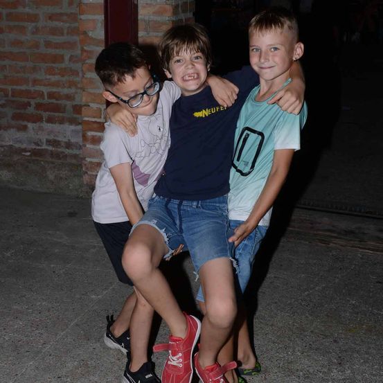 Drei Jungen stehen eng beieinander draußen und lächeln. Ein Junge trägt eine Brille und hält die Schultern des Jungen vor ihm.