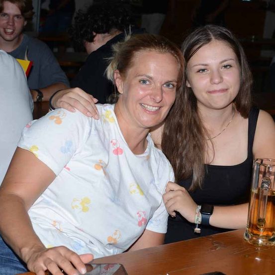 Zwei Frauen sitzen an einem Tisch und lächeln, eine hält ein Glas Bier. Hinter ihnen sitzen andere an einer Bank, ein Mann lacht.