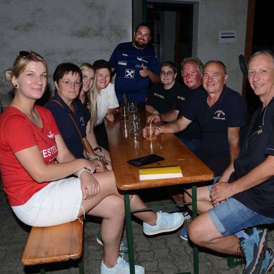 Eine Gruppe von Menschen in passenden Shirts sitzt an einem Tisch. Sie haben Gläser und ein Handy auf dem Tisch. Sie scheinen für ein Foto zu posieren.