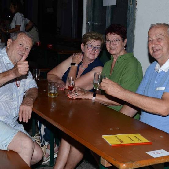 Vier ältere Erwachsene sitzen an einem Tisch, lächeln und halten Gläser mit Wein. Der Mann auf der linken Seite gibt einen Daumen hoch. Der Tisch hat eine gelbe Menükarte.